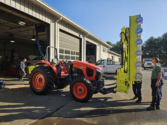 A large red tractor sitting outside of a garage bay with a yellow machine loaded on the front. 
